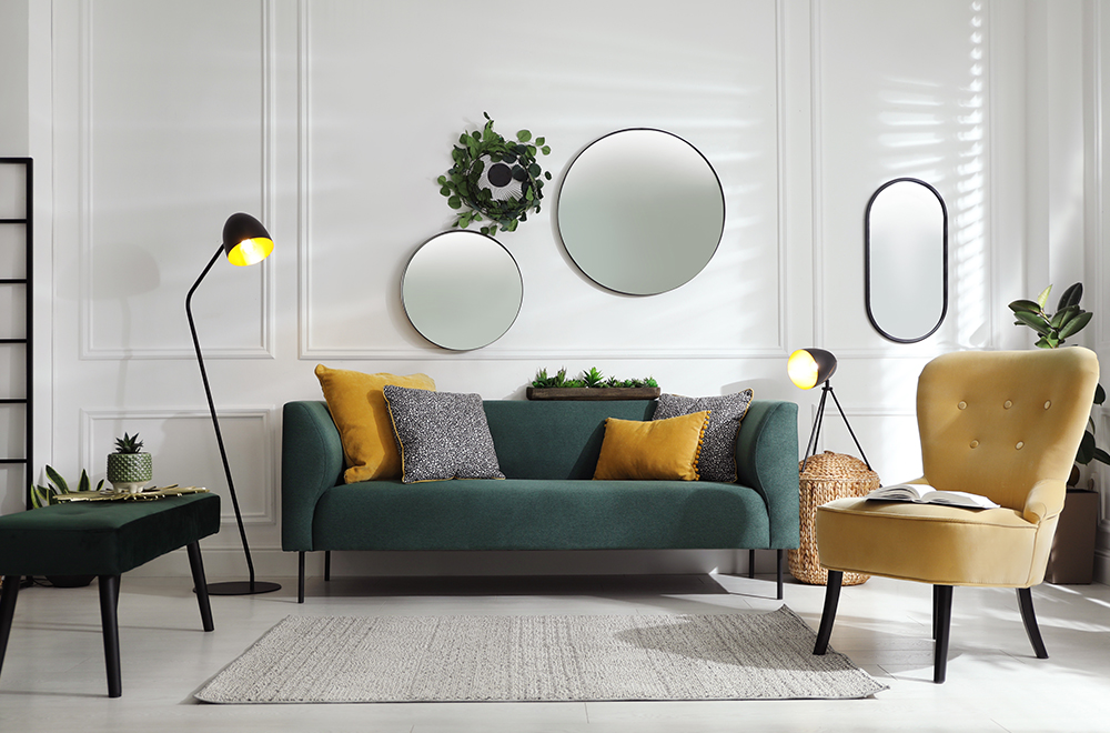 Moderní obývací pokoj ve světlých barvách se zelenou pohovkou, žlutými polštáři a kulatými zrcadly na zdi.