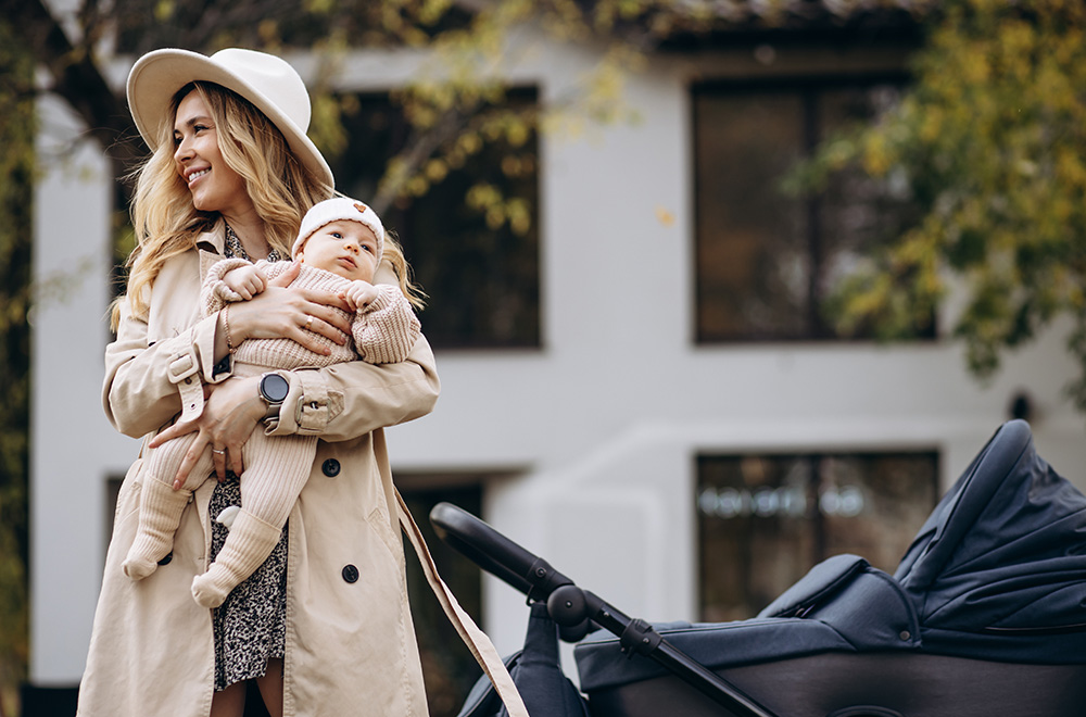 Žena drží v náručí dítě a jde vedle kočárku před budovou.
